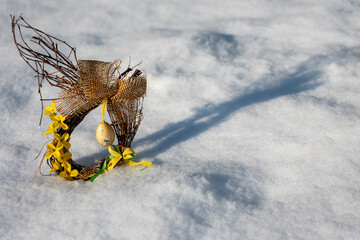Easter wreath made of twigs with decorative egg and yellow flowers placed on snow outdoors. Spring holiday decoration symbolizing the transition from winter to Easter season.