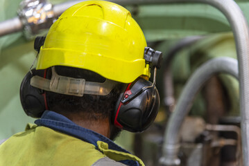 Back view of marine engineer wearing protective helmet and hearing protection while inspecting main engine in ship engine room.