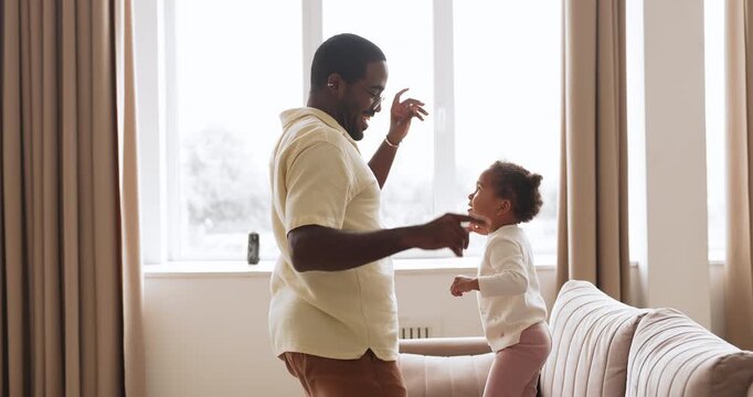 Active toddler biracial girl holding loving black father hands and jumping on couch, happy family dancing, having fun together in living room at home. Play time with children, bonding and fatherhood