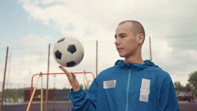 caucasian freestyler balancing football on hand at playground, blue windbreaker and calm expression, swings and poles in background, skillful spin and controlled juggle, youthful confidence, soft