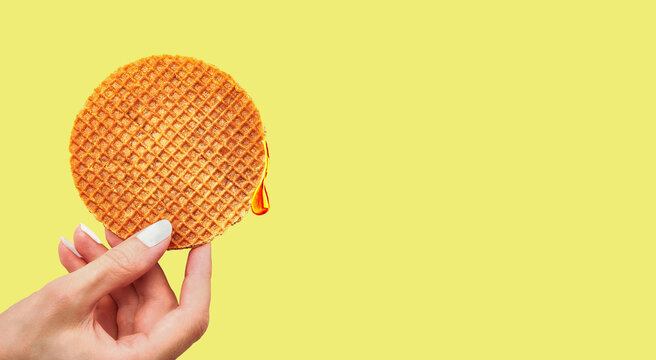 Female's hand holding stroopwafel with caramel drop over yellow background. Traditional Dutch waffle caramel cookie. Delicious snack concept, closeup shot, horizontal orientation, copy space