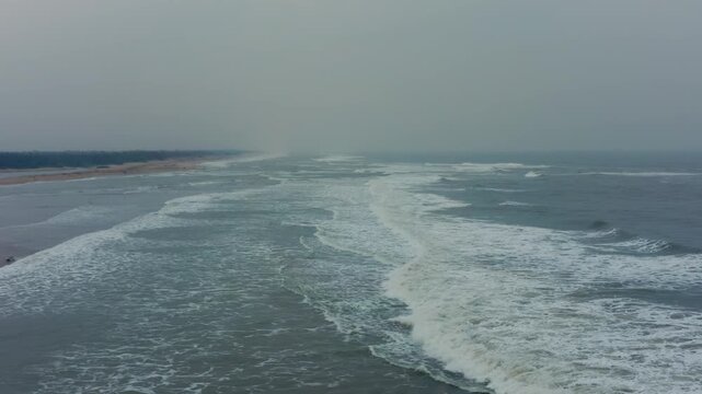 Sweeping drone aerial tracks continuous waves rolling along Chandrabhaga Beach's hazy shoreline near Konark Sun Temple in Odisha, India.