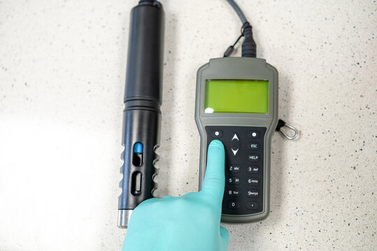 Female hands in protective gloves adjusting settings on a handheld pH meter.