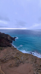 Fototapeta premium A scenic view of a rocky coastline with turquoise waters under a cloudy sky. Waves crash against the shore, creating a serene atmosphere.