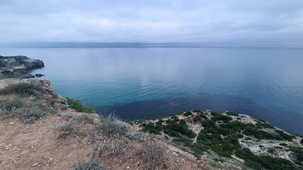 Fototapeta premium A serene coastal landscape featuring a rocky cliff overlooking calm blue waters. The sky is overcast, creating a tranquil atmosphere.