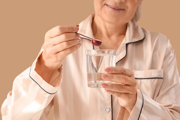 Mature woman in pajamas taking collagen powder on beige background, closeup