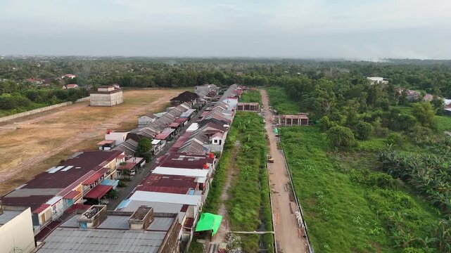 A drone shot from the air captures a long row of townhouses along a quiet residential street, surrounded by green fields and undeveloped land.
