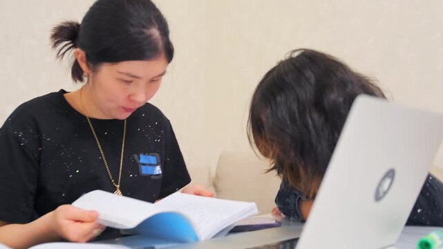 mother helping daughter learn at laptop, guiding daughter through workbook during focused study session, asian mother patiently assisting daughter with homework beside laptop in calm setting