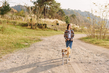 Woman walking a dog along a dirt path in a countryside setting, casual plaid shirt and jeans, serene outdoor scene with trees and soft sunlight, daily activity and companionship.