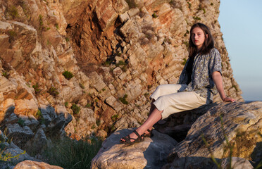 Naklejka premium A girl poses by the sea, standing on rocks near the slope of a high cliff, panoramic view of the seashore and mountains at sunset