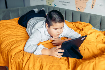 A young Asian boy with short black hair lies on a bed, focused on a tablet. The bed is covered with an orange blanket, and a map is visible on the wall.