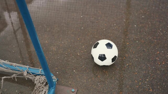 soccer ball near wet goalpost, puddles empty urban pitch with glistening asphalt, blue metal post and frayed net, closeup of ball rolling after missed kick, overcast sky and reflective puddles