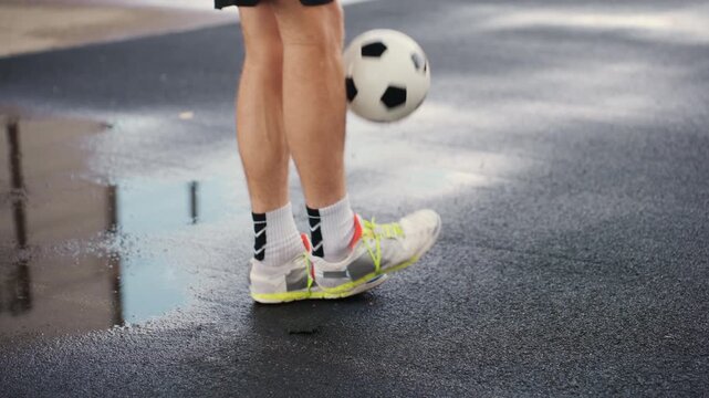 male legs juggling soccer ball rainy asphalt, closeup training routine with neon sneakers and ankle socks, puddles and reflective surface, controlled touches and light splashes, rhythmic footwork