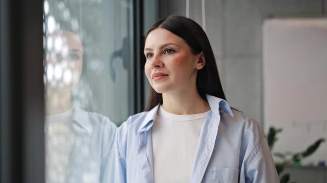 Thoughtful woman gazes out window, displaying calm and reflective demeanor. Contemplative moment in modern office space, expressing quiet hope and serenity.