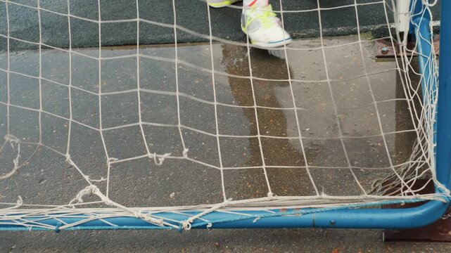 Wet goal net with soccer ball on rainslick urban pitch, puddles reflecting net and shoe prints, youth striker steps back after shot, ball settles inside net then rolls away, damp sneakers leaving
