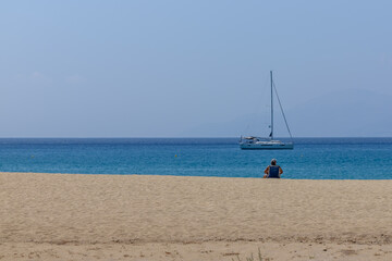View of a person sitting at the beach enjoying the view and a sailboat in the distance in Greece