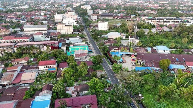 The aerial drone footage shows a bustling suburb with rows of densely packed houses, a main road busy with construction and buildings under a clear sky.