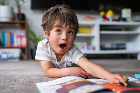 A young boy lies on a soft carpet, amazed as he reaches for a book in a bright living room.