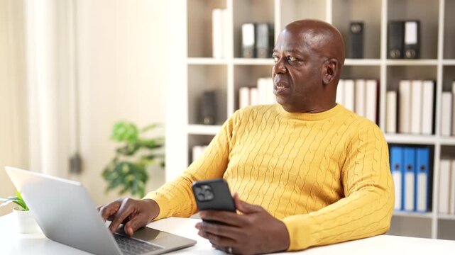 Worried mature african american businessman feeling stressed while multitasking on his smartphone and laptop, showing frustration and anxiety from receiving bad news at his office desk
