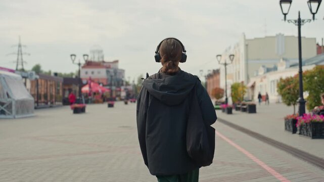 woman walking away along promenade rear view captures introspective journey with hoodie and backpack silhouette against empty boulevard lined with planters and storefronts under grey sky headphones