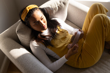 Mid-adult Asian woman reclining in armchair checking pink headset in mustard overalls near window