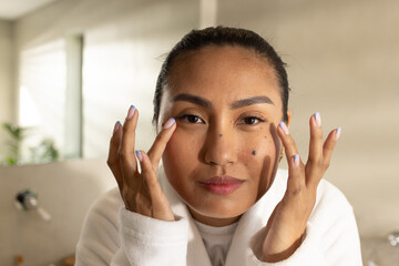 Mid adult Asian woman massaging under eyes at bathroom sink wearing white robe near frosted window