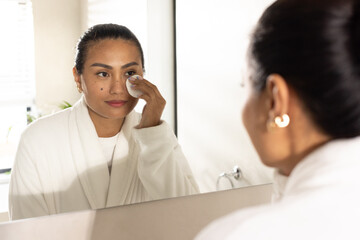 Mid adult Asian woman wearing robe standing at bathroom mirror, removing makeup with cotton pad