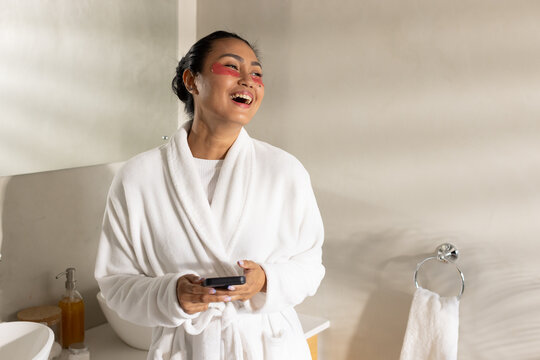 Asian woman laughing while holding smartphone at bathroom vanity wearing white terry bathrobe