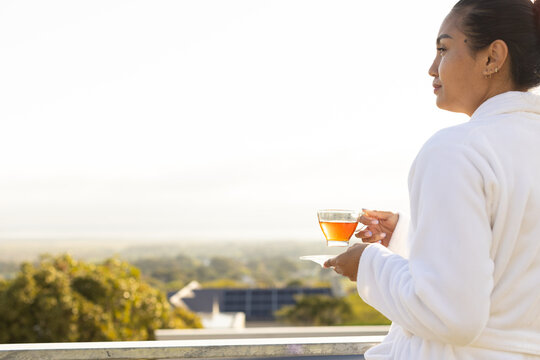 Asian woman standing on balcony in robe holding clear glass teacup on white saucer, copy space