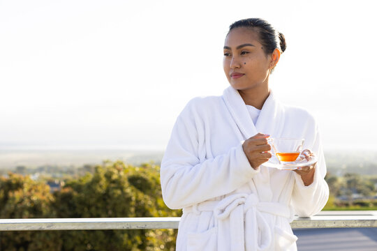 Asian woman in white bathrobe standing holding teacup and saucer by balcony railing, copy space