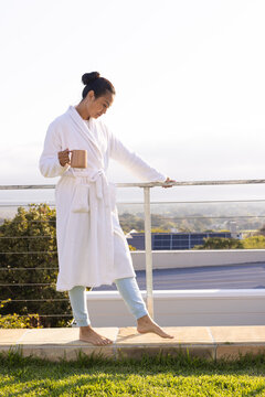 Asian woman wearing white bathrobe balancing on balcony ledge holding brown mug using metal railing