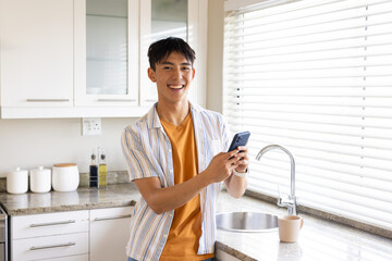 Asian man standing at kitchen counter smiling, holding phone wearing orange tee near sink mug