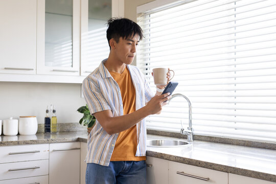 Adult Asian man leaning at kitchen counter, holding ceramic mug and checking phone in striped shirt
