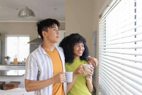 Diverse couple leaning in kitchen near blinds, in green top and striped shirt holding mugs
