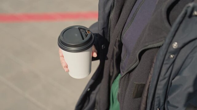 stylish streetwalker holding paper coffee cup, navy parka layered over green sweater, midday sunlight on sidewalk, closeup focus on cup and hand, relaxed stride near red curb, fashionforward casual