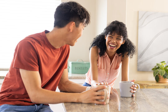 Couple in casual clothing sitting at kitchen counter holding mugs and laughing by biscuit tin
