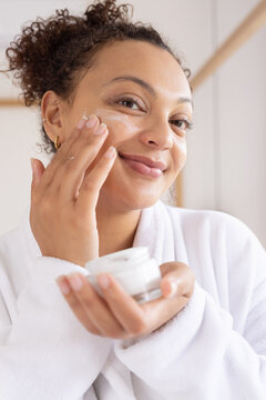African American woman using cream with tips holding jar wearing white robe gold hoops in bathroom