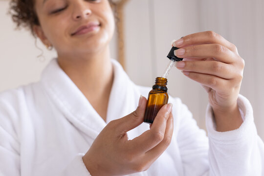 African American woman in robe holding amber dropper bottle, lifting pipette, showing nails at sink