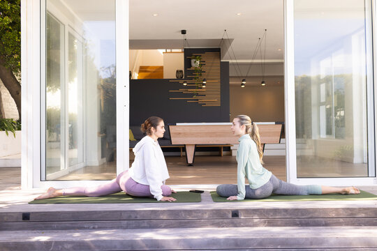 Female friends sitting on yoga mats on wood deck, stretching in leggings with smartphone between