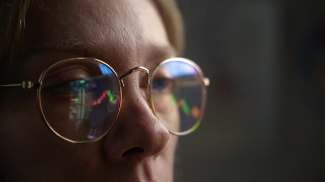 Close up of a woman trader watching scree, glasses reflecting financial charts, long blond hair and fresh natural face, working at a desktop with screen glow in dim, shadowy lighting.