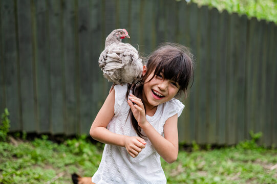 Girl laughs joyfully while playing with a chicken in a backyard with green grass and fence