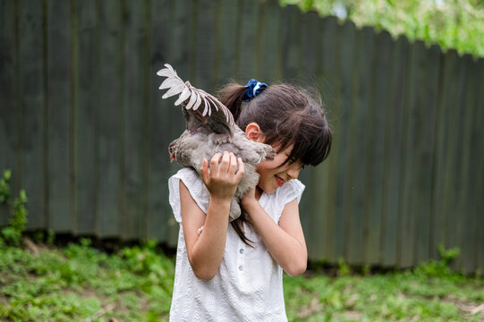 Girl aged six to eight smiles while holding a chicken in a green backyard