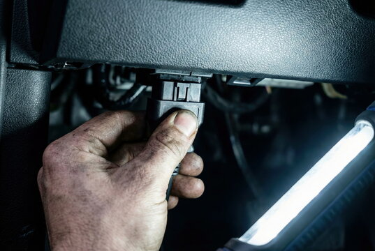 Dirty mechanic's hand connects an OBD-II diagnostic scanner to a car's port under the dashboard, illuminated by a work light, for vehicle repair and maintenance.