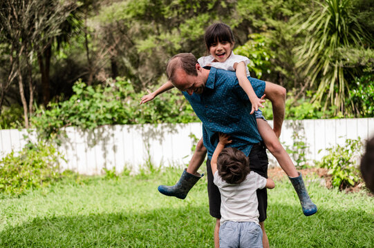 A man enjoys a fun moment lifting one child while another child tugs at his waist in a garden