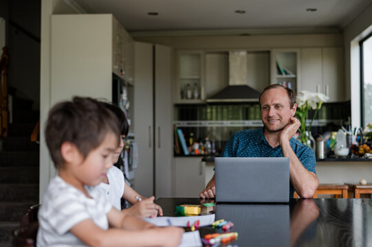 A man watches his children draw and create art at the kitchen table in their home