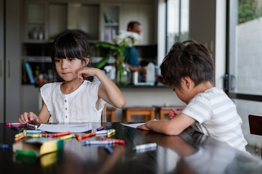 Children focus on their colouring activities at a kitchen table with an adult in the background