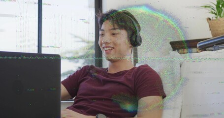 Smiling maroon-tee Korean teen leaning on couch in lounge, wearing black headphones watching laptop © vectorfusionart