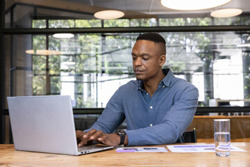 African american man typing on laptop at office desk with charts and glass of water