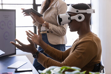 Diverse coworkers interacting using vr headset, tablet at office desk near monitor