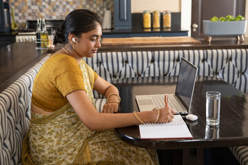 Indian woman writing in notebook at home kitchen nook and in yellow floral sari with laptop © wavebreak3